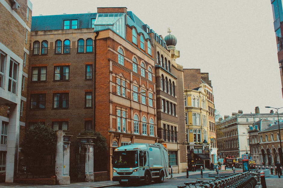 A street scene in an urban area featuring a row of old, multi-story buildings with Victorian-style architecture, characterized by brick facades, arched windows, and decorative trim. The buildings are situated along a paved sidewalk with bollards and bike racks visible in the foreground. A medium-sized, light blue waste collection truck is parked on the street close to a gated entrance with stone pillars, which may indicate a private property or property boundary. The truck's surface reflects nearby structures and ambient light. In the background, there are additional historic-style buildings, a lamppost, and a bus or large vehicle further down the street. The overall atmosphere suggests a busy city environment, with a neutral daylight lighting that highlights the textures of the brickwork and architectural details, aligning with rubbish removal services or private waste collection common in such urban settings.