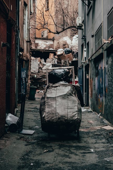 A narrow alleyway filled with overflowing rubbish and discarded materials, featuring a large, grey, industrial-style trash bag in the foreground, which appears to be heavily filled and slightly dirty. Behind the bag, there are stacks of cardboard boxes, paper, and plastic waste piled haphazardly against a wooden barrier at the end of the alley, indicating an area used for private rubbish disposal or collection. The alley is flanked by aged buildings with brick and concrete facades; one side has graffiti on the walls, and the other side has metal window grilles and pipes attached to the building exterior. The ground is uneven with patches of dirt and spilled debris, and the lighting suggests an overcast day or shaded environment, emphasizing the cluttered and neglected appearance typical of a location awaiting rubbish clearance services. The scene conveys a sense of an independent collection point or an area used for temporary waste storage near urban residential or commercial buildings, aligning with alternative rubbish removal options.