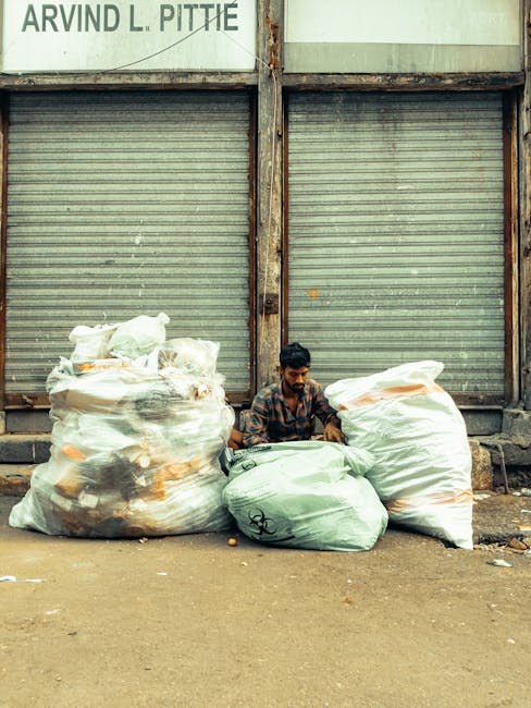 A man with dark hair and a beard, wearing a patterned shirt, is seated on the ground between two large white plastic bags filled with mixed waste materials in front of a closed, grey, corrugated metal shutter. To the left of him, the first bag contains various rubbish items, including plastic bottles and paper, with the fabric looking slightly crumpled and transparent enough to reveal its contents. On his right, a second bag with a similar appearance holds additional waste, and a third, smaller bag is positioned directly in front of him, also filled with debris. Above the scene, a weathered sign with partially visible text, including the name “ARVIND L. PITTIE,” is mounted on the wall above the shutter, indicating a commercial or industrial setting. The environment appears to be an outdoor urban area with a concrete or asphalt ground, showing signs of dirt and scattered litter, characteristic of an area requesting independent waste collection solutions as part of alternative rubbish removal practices. The overall scene, captured in natural light, reflects a context where informal waste management is taking place outside a closed storefront or storage unit, consistent with private rubbish handling methods. 