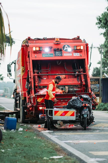 A waste collection worker wearing a high-visibility yellow and red uniform is operating a large red rubbish and waste collection truck, positioned on the side of a road. The worker is seen leaning over a small metal container, possibly preparing to load or unload waste materials. The back of the truck has an open panel revealing internal mechanisms, with visible hydraulic components and safety markings, including a visible license plate reading 'BRJ 7751'. In front of the worker, a motorcycle with a black plastic bag secured on its seat is parked, indicating on-site waste handling. The surrounding environment features a partially visible sidewalk, with some litter and debris near the curb, and a street scene in the background with trees, electrical wires, and distant signage, suggesting a suburban or urban area. Overcast daylight provides diffuse lighting, emphasizing the industrial and practical context of rubbish collection, aligned with alternative waste management practices employed by independent waste services such as those offered by wastecollectionelephantandcastle.co.uk.