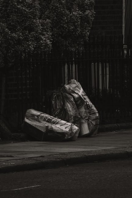 A man with dark hair and a beard, wearing a patterned shirt, is seated on the ground between two large white plastic bags filled with mixed waste materials in front of a closed, grey, corrugated metal shutter. To the left of him, the first bag contains various rubbish items, including plastic bottles and paper, with the fabric looking slightly crumpled and transparent enough to reveal its contents. On his right, a second bag with a similar appearance holds additional waste, and a third, smaller bag is positioned directly in front of him, also filled with debris. Above the scene, a weathered sign with partially visible text, including the name “ARVIND L. PITTIE,” is mounted on the wall above the shutter, indicating a commercial or industrial setting. The environment appears to be an outdoor urban area with a concrete or asphalt ground, showing signs of dirt and scattered litter, characteristic of an area requesting independent waste collection solutions as part of alternative rubbish removal practices. The overall scene, captured in natural light, reflects a context where informal waste management is taking place outside a closed storefront or storage unit, consistent with private rubbish handling methods. 
