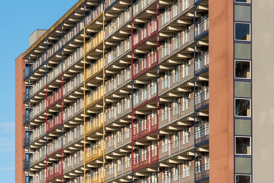 A high-rise residential building features multiple levels of small balconies arranged in a uniform grid pattern. Each balcony has a metal railing painted in alternating colors of red, yellow, and blue, creating a vibrant, geometric appearance. The balconies are made of concrete with a textured finish, displaying a neutral light gray tone. The building's exterior wall, visible on the right, is composed of large, square windows set into a smooth, light beige facade, reflecting the sky and surrounding environment. The image is captured in natural daylight, highlighting the sharp lines and bold colors of the balcony railings against the neutral building surface. The scene exemplifies modern urban architecture, with balconies that may be used for personal outdoor space or storage, aligning with contemporary housing layouts. This type of building exemplifies how private outdoor areas are integrated into high-density living environments, sometimes requiring external rubbish removal services for maintenance or clearance of accumulated debris on communal balconies or estate grounds, which companies like wastecollectionelephantandcastle.co.uk can assist with.