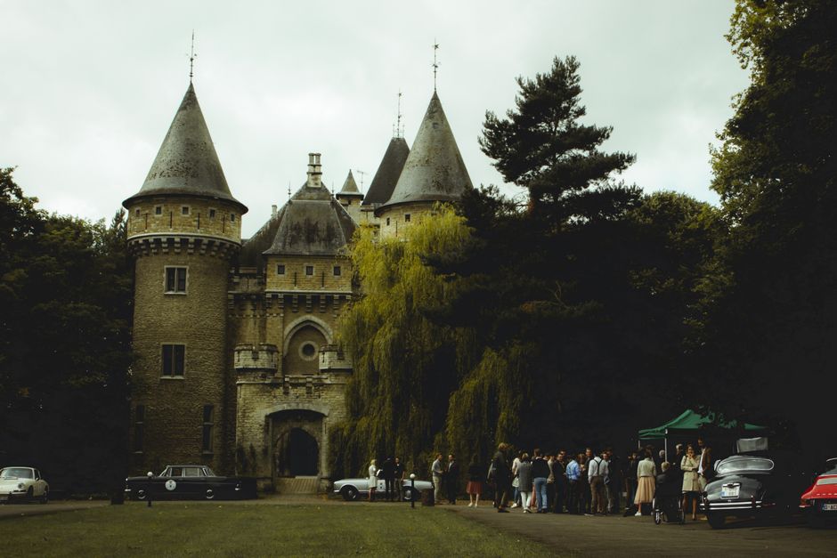 A stately historic building with a symmetrical façade featuring multiple large rectangular windows, painted in a pale yellow or beige tone with white trim. The roof is dark grey, and behind it rises a small bell tower with a greenish spire. In the foreground, a gravel pathway leads directly towards the building, flanked by neatly maintained grass lawns on either side. Centered along the path is a decorated Christmas tree adorned with small white or warm yellow lights, surrounded by small, rounded shrubbery. To the right, an adjacent brick church or chapel with a steeply pitched dark roof and a prominent tower with a spire is visible, with some bare trees in the background suggesting it is late autumn or early winter. The scene is lit by soft, natural daylight with a slightly overcast sky, creating a calm and orderly outdoor atmosphere typical of a historic estate estate or public park, possibly within a private property managed for regular visits. No visible waste materials or rubbish are present in the scene, aligning with professional site maintenance and clean management practices as part of independent waste handling or estate upkeep.