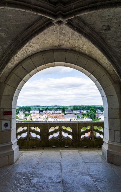 The image shows a view through a large, rounded stone archway with a smooth, lightly weathered texture. The arch is part of a historic building or structure, with detailed stonework and a polished stone floor visible at the base. Beyond the arch, there is a stone balustrade with decorative, curved balusters, painted in a muted yellow or beige tone. The balustrade provides a partial view over a townscape consisting of white and pastel-colored buildings with grey roofs, spread out across a flat landscape under a clear blue sky with some wispy clouds. The scene suggests an elevated position, possibly a tower or castle, overlooking a suburban or small urban area. The lighting indicates daylight, with natural light illuminating the stone surfaces and casting soft shadows. In context, this view might relate to the importance of proper waste management within historic or privately owned properties, emphasizing the need for specialised rubbish removal services suited to such buildings.