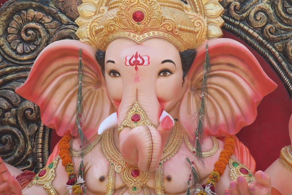 Close-up image of a large, elaborately decorated statue of the Hindu deity Ganesha, featuring a pink and cream-colored elephant head with detailed facial features, including large ears, expressive eyes, and a prominent trunk. The statue is adorned with intricate gold jewelry, including a crown embedded with a red gemstone and ornate necklaces, as well as a decorative forehead mark. The surrounding background displays a detailed, traditional patterned backdrop with swirling designs in gold, black, and red tones, emphasizing the cultural and spiritual significance of the sculpture. The texture of the statue's surface appears smooth with a polished finish. Fresh flower garlands and small decorative accessories are placed around the statue, enhancing its presentation for religious or ceremonial purposes. The lighting in the image highlights the vibrant colors and intricate details of the statue, creating a visually striking focal point that reflects cultural reverence and artistry within a spiritual context. This detailed visual description aligns with the practice of documenting religious statues for informational and accessibility purposes, supporting understanding of cultural heritage materials relevant to waste collection and disposal environments involving religious or ceremonial artifacts.