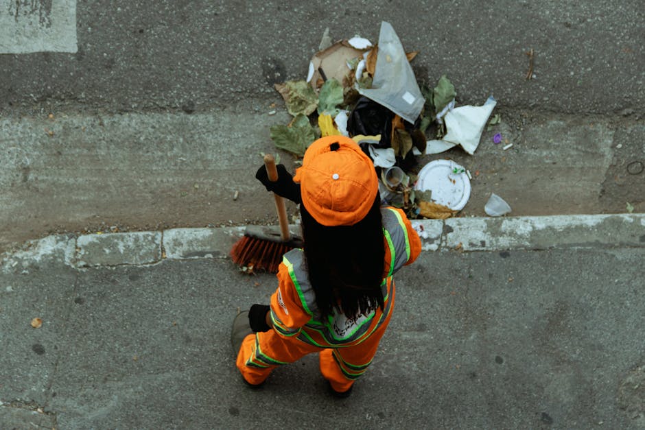 An individual wearing a high-visibility orange jacket and matching hat, seen from above, is collecting and clearing litter from the edge of a paved sidewalk and road. The person is holding a broom and appears to be engaged in rubbish removal work. Scattered on the ground near the curb are various waste items, including crumpled paper, plastic packaging, a disposable cup, and dried leaves. The surface of the sidewalk is textured concrete, with the curb separating it from an asphalt road that shows signs of wear. The scene suggests a focus on clearing public space rubbish, possibly as part of an on-site clearance service conducted by a waste collection company. The environment is well-lit, and the backdrop includes a rough-textured concrete wall or pavement edge. This visual context aligns with independent waste handling or private rubbish removal, providing a clear depiction of a tidy-up operation in an urban setting.