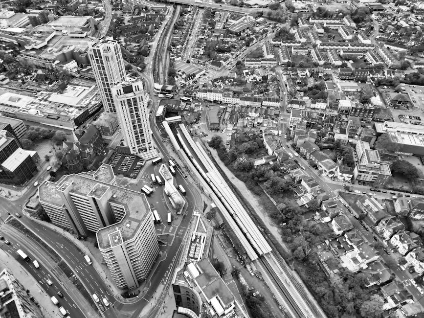 An aerial view of a dense urban area in black and white, showing high-rise office buildings with flat roofs and glass facades in the foreground. The streets are filled with multiple vehicles, including buses and cars, moving along curved roadways that surround the buildings. To the right, a railway track runs diagonally across the scene, with a station platform visible near the center of the image. The railway line is bordered by trees and residential houses with pitched roofs, closely packed together, indicating a mixed-use neighbourhood. The background reveals a network of roads, smaller buildings, and scattered greenery, all captured under diffused daylight, highlighting the textures and contrasts of the cityscape. The scene illustrates a typical city centre environment where private and commercial transportation infrastructure coexist with densely packed housing, aligning with themes of urban waste management and alternative collection methods that a company like wastecollectionelephantandcastle.co.uk might serve in managing rubbish removal and disposal in busy metropolitan areas.