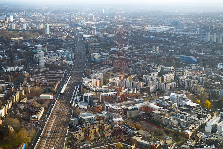 An aerial view of a dense urban area in black and white, showing high-rise office buildings with flat roofs and glass facades in the foreground. The streets are filled with multiple vehicles, including buses and cars, moving along curved roadways that surround the buildings. To the right, a railway track runs diagonally across the scene, with a station platform visible near the center of the image. The railway line is bordered by trees and residential houses with pitched roofs, closely packed together, indicating a mixed-use neighbourhood. The background reveals a network of roads, smaller buildings, and scattered greenery, all captured under diffused daylight, highlighting the textures and contrasts of the cityscape. The scene illustrates a typical city centre environment where private and commercial transportation infrastructure coexist with densely packed housing, aligning with themes of urban waste management and alternative collection methods that a company like wastecollectionelephantandcastle.co.uk might serve in managing rubbish removal and disposal in busy metropolitan areas.